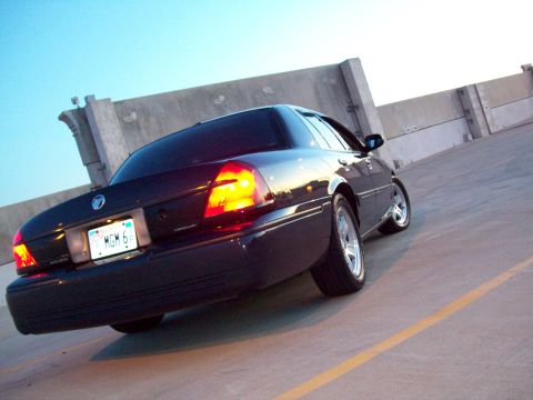 1999 Mercury Grand Marquis - The rear of a dark sedan is captured from a low angle against a concrete wall and clear sky.