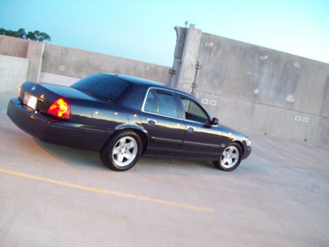 1999 Mercury Grand Marquis - A dark sedan is parked outdoors near a concrete wall under a clear sky.
