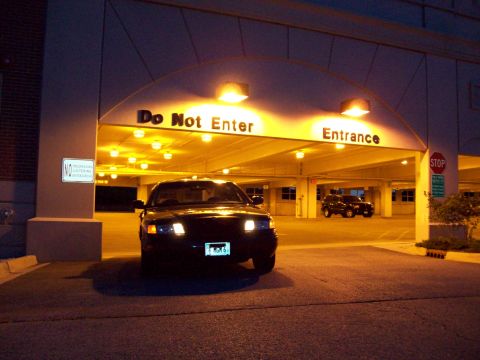 1999 Mercury Grand Marquis - A dark Ford Crown Victoria is parked in front of a parking garage entrance with 'Do Not Enter' and 'Entrance' signs illuminated.