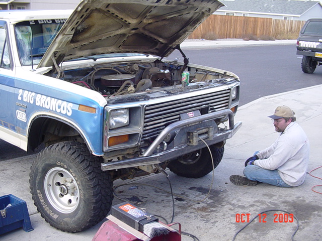 Man servicing a blue and white full-size Ford Bronco with hood up