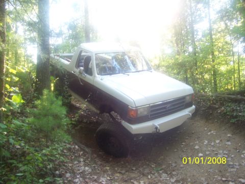 1989 Ford Bronco - A two-tone Ford Bronco navigates a wooded trail with a modified front bumper.