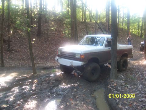 1989 Ford Bronco - A lifted Ford pickup truck navigates a muddy trail in a wooded area, with another vehicle in the distance.