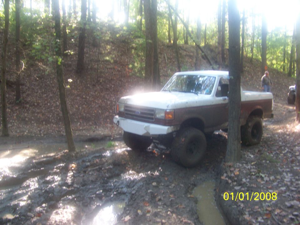Two-tone Ford pickup truck with aftermarket bumper driving through mud in a forest. Another vehicle is visible in the background
