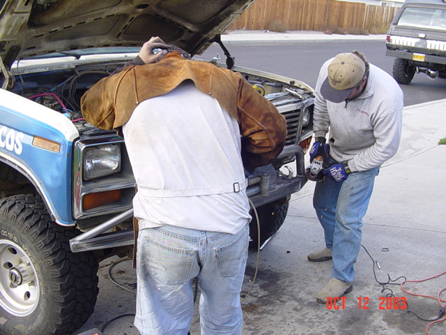 Two men in welding gear working at the front of a blue Ford Bronco on a driveway, trucks behind, Oct 2003