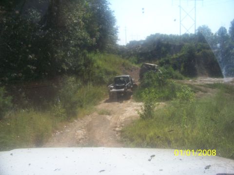 1989 Ford Bronco - Two off-road vehicles navigate a dirt path surrounded by lush greenery under a bright sky.