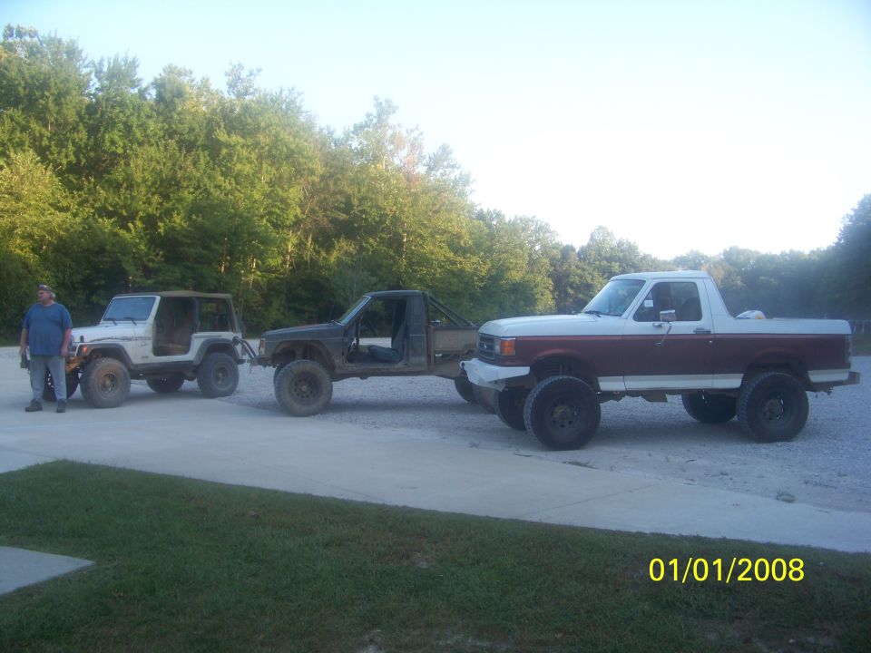 Two-tone red and white pickup truck with lifted suspension and large tires, towing a modified Jeep