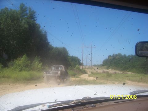 1989 Ford Bronco - Two trucks driving on a dusty dirt road with power lines in the background.