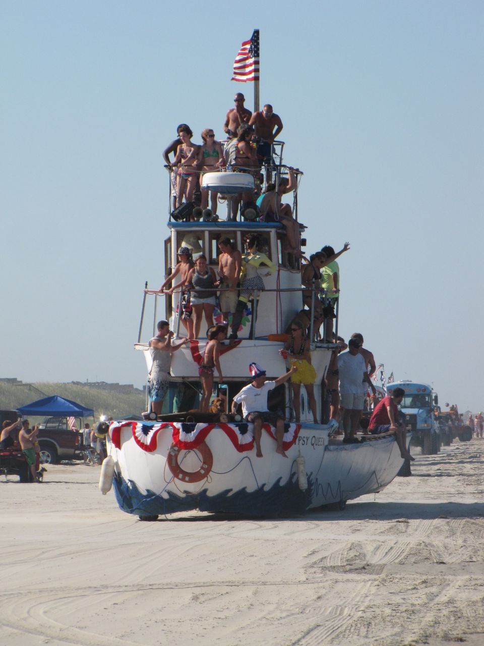 Boat named Gypsy Queen on a beach with many people on its decks, American flag flying