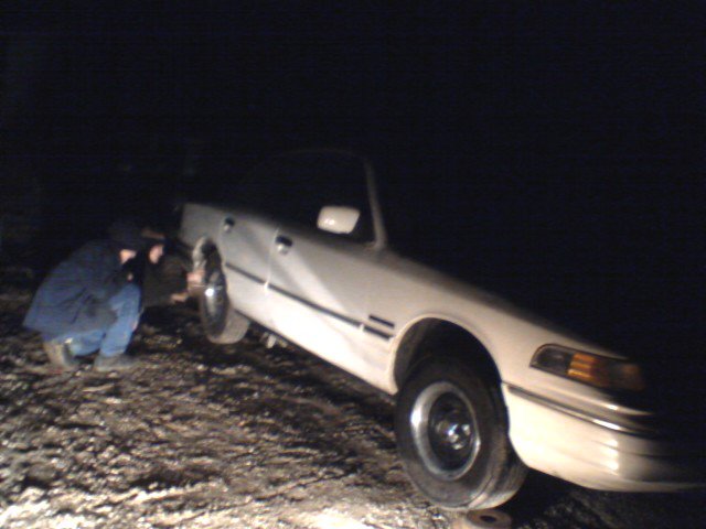 White sedan with two people working on the front wheel. The car is on uneven ground at night