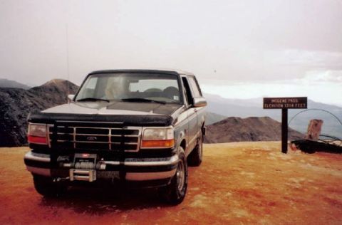 1995 Ford Bronco - A dark green/black full-size Ford Bronco with a black bra over the hood and grille, a front-mounted winch and a whip antenna, parked on the…