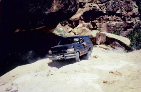 1995 Ford Bronco - A black SUV navigates a dusty, rocky terrain under a large rock overhang.