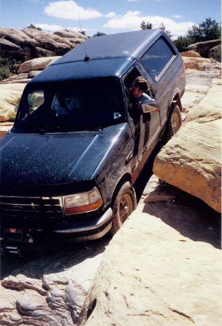 1995 Ford Bronco - A dark green full-size Ford Bronco straddling a slickrock crack in Utah red-rock country, front driver wheel dropped into the gap while the…