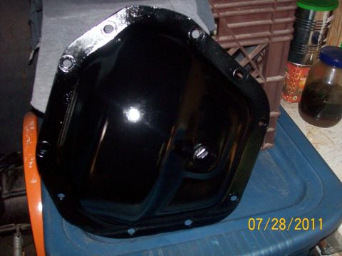 1982 Ford F-100 - A gloss-black stamped-steel differential cover with ten bolt holes and a threaded fill plug, sitting face-up on a blue plastic tote in a sh…