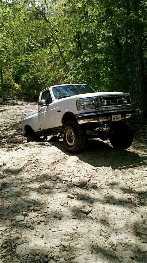 1995 Ford F-150 - White 1995 Ford F-150 lifted truck driving on a dirt trail through a wooded area.