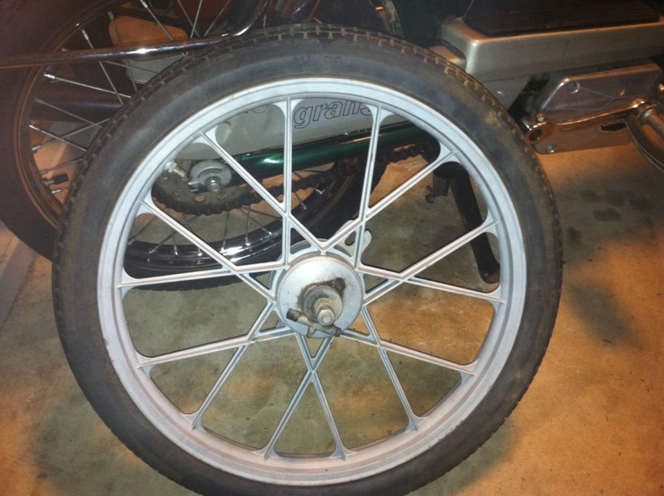 Close-up of a silver 10-spoke motorcycle wheel with a tire. Text 'grana' is visible on the rim. Another wheel is behind it