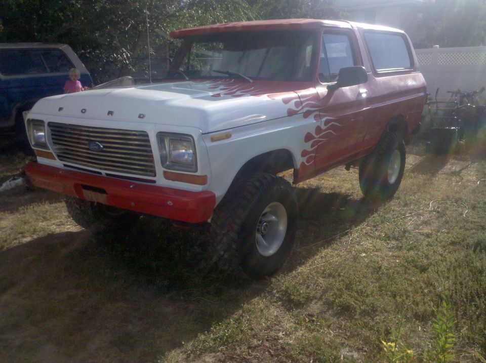 Red and white Ford Bronco with flame decals, lifted on large tires, parked on grass