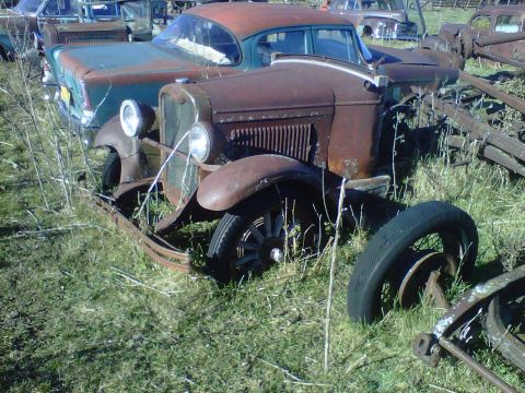 1978 Chevrolet C/K Pickup - A rusty brown vintage car sits in a junkyard, with a teal car partially visible behind it.