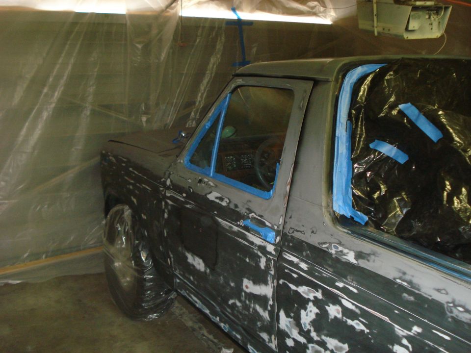 Pickup truck body covered in primer and blue masking tape, inside a plastic-covered booth