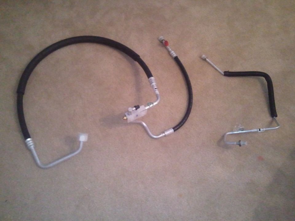 Three automotive AC hoses laid out on a carpeted surface. One large curved hose with a white fitting, and two smaller straight hoses
