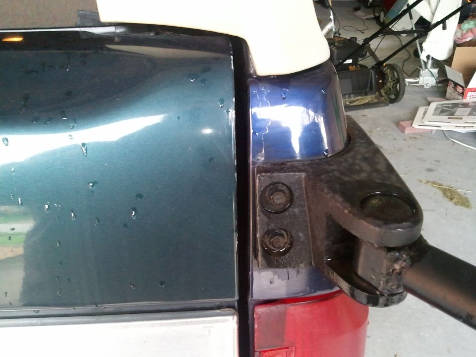 Close-up of rear corner of blue SUV, showing part of the tailgate and a black bracket. Water droplets on the paint