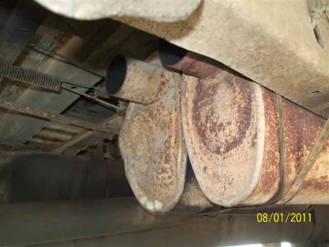 Rusty muffler and exhaust components viewed from under a truck, with brake cable and frame visible