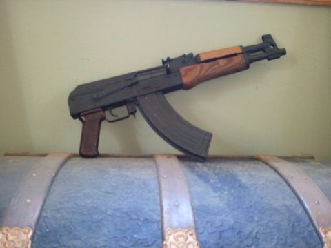 1985 Ford F-150 - An AK-47 style rifle with wooden furniture rests on a decorative blue chest. The image includes a 'SUPERMOTORS' watermark.