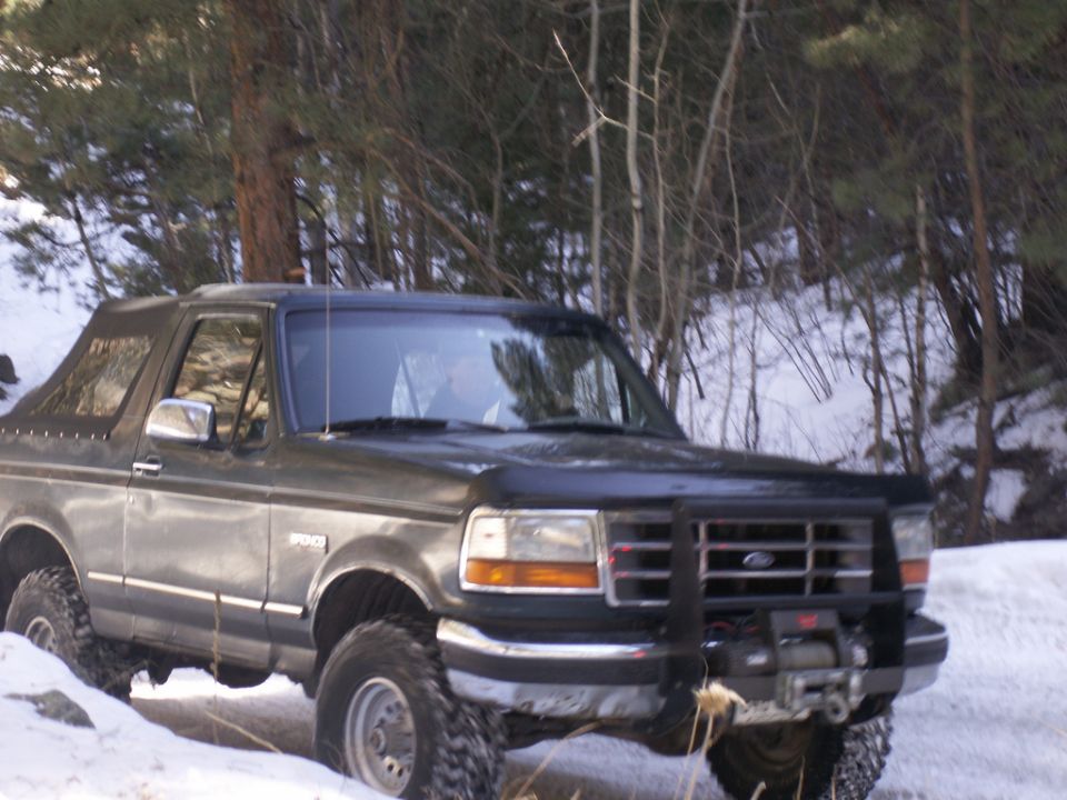 Dark gray Ford Bronco with a winch on the front bumper, angled from the front-right in a snowy wooded area