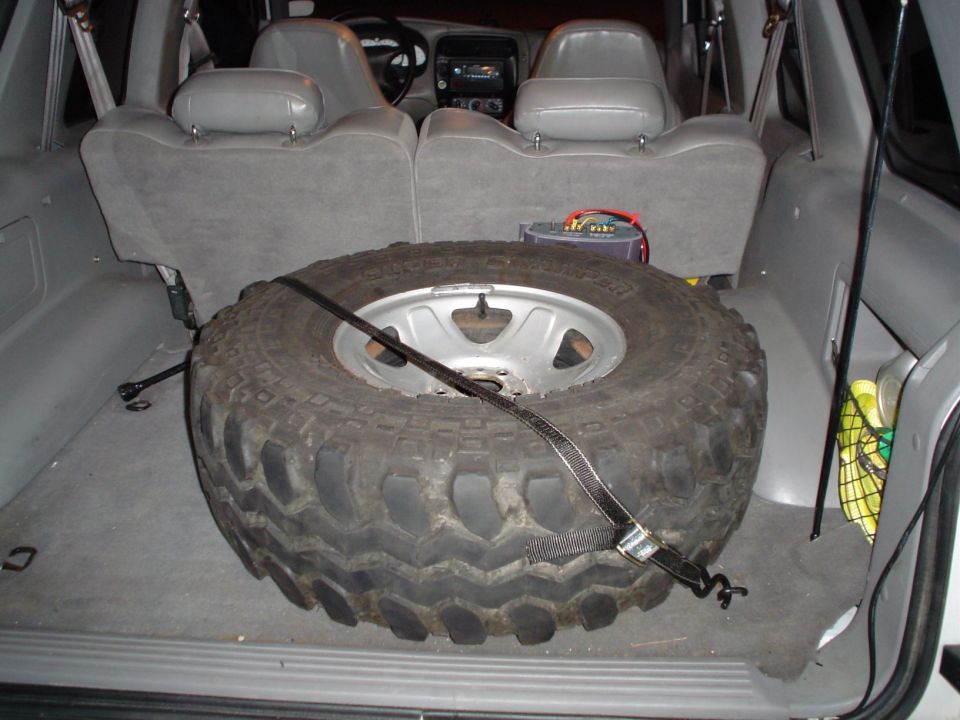 Muddy spare tire on silver wheel strapped down in gray SUV cargo area