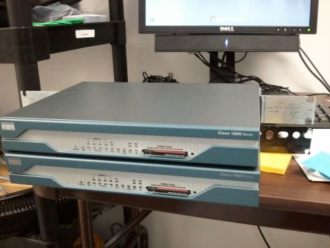 1985 Ford F-150 - Two Cisco 1800 Series network routers are stacked on a wooden desk next to a computer monitor.