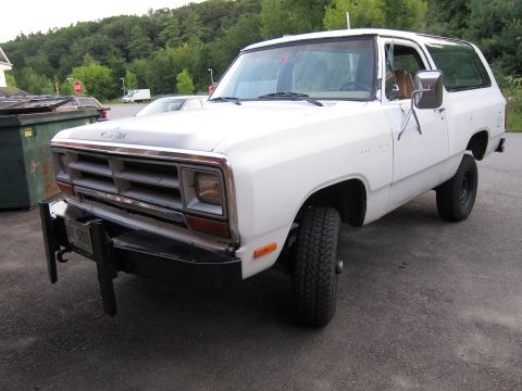 1987 Dodge 1500 - A white SUV is parked on asphalt with trees and dumpsters in the background.