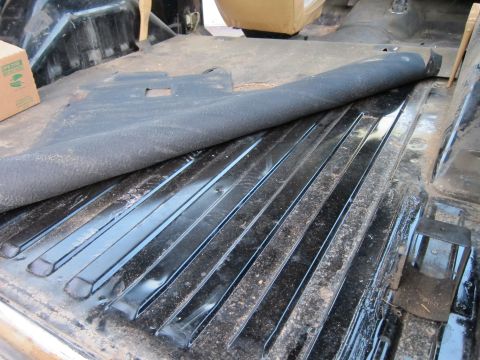 1987 Dodge 1500 - The interior floor of a vehicle shows exposed ribbed metal and a partially lifted mat, with a cardboard box in the background.