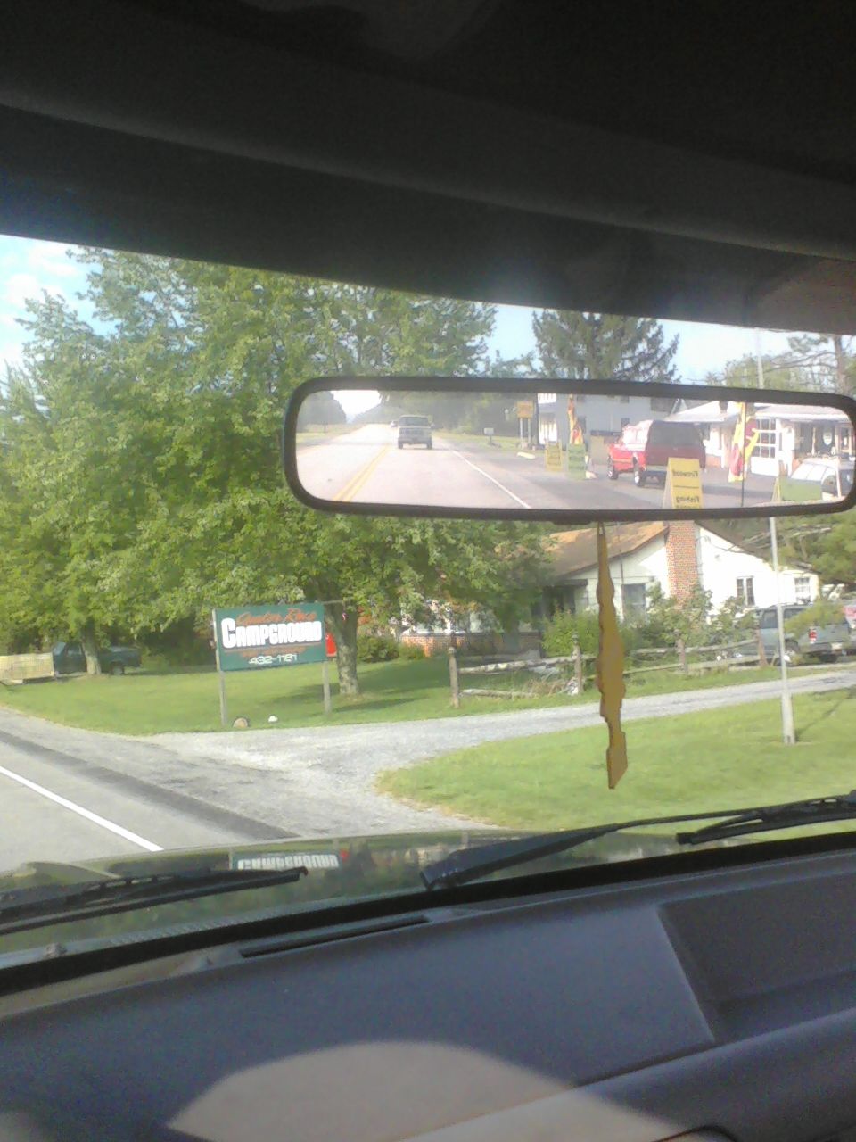 View from inside a vehicle, looking out the windshield and through the rearview mirror at a road with trees and houses. A red van and another vehicle are visible in the mirror