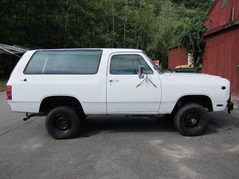 1987 Dodge 1500 - A white SUV with a camper shell is parked outdoors next to a red building.