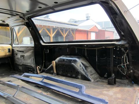 1987 Dodge 1500 - The interior of a truck cab is shown with the doors removed and the floor stripped bare, revealing the wheel wells and wiring.