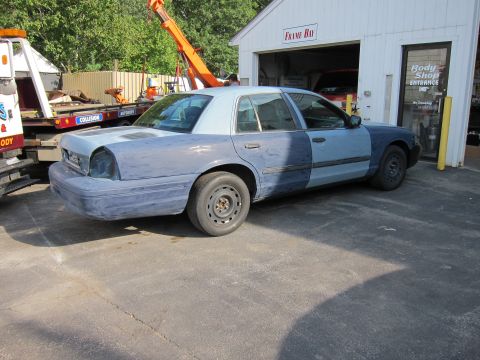 1987 Dodge 1500 - A sedan in primer, with two-tone blue doors, sits outside a body shop, awaiting repairs.