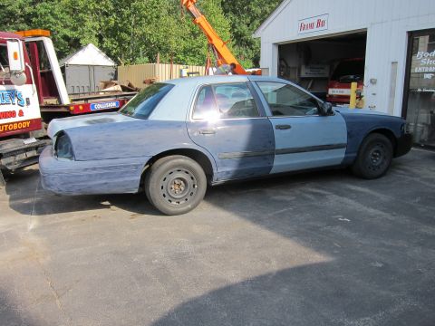 1987 Dodge 1500 - A blue sedan is parked outside a body shop, showing primer on its rear quarter panel and front fender.