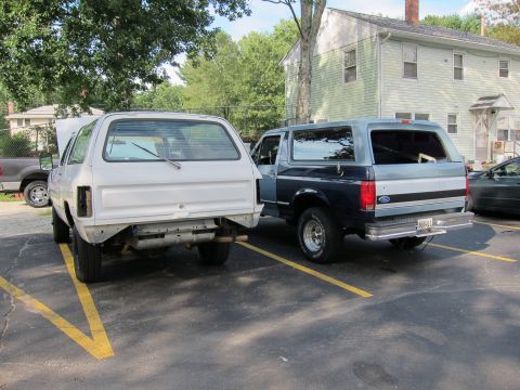 1987 Dodge 1500 - Two SUVs are parked in a lot, one white and appearing to be undergoing repairs, the other a two-tone blue and grey.