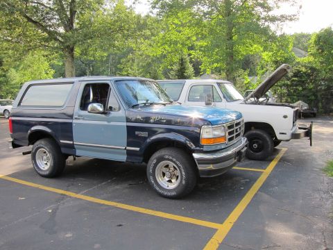 1987 Dodge 1500 - A two-tone Ford Bronco is parked next to a white pickup truck. The Bronco appears to be from the early to mid-1990s.