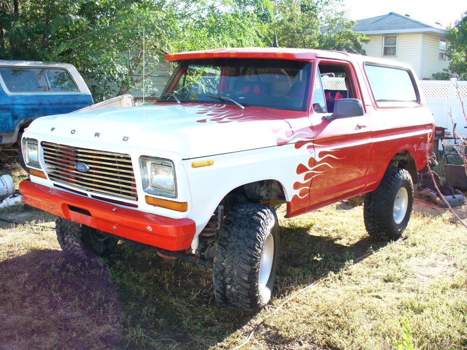 White and red Ford Bronco with flame decals, lifted with big tires. Another blue Bronco is parked behind it