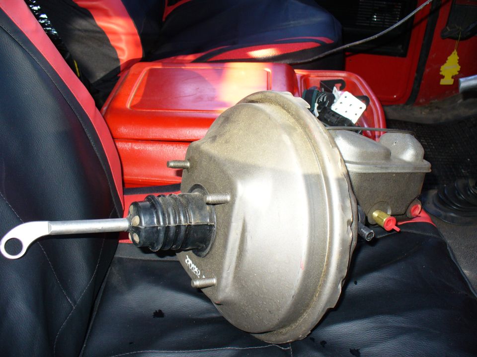 Brake booster and master cylinder assembly on black and red seats, with a red plastic container behind it