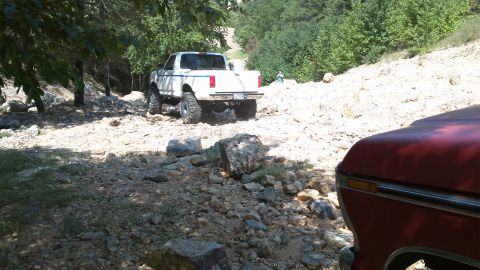 1978 Ford Bronco - A white pickup truck navigates a rough, rocky path with trees and foliage surrounding the area.