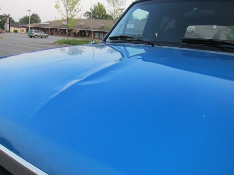 2001 Ford Crown Victoria - A blue vehicle's hood shows a significant dent, with a silver minivan visible in the background parking lot.