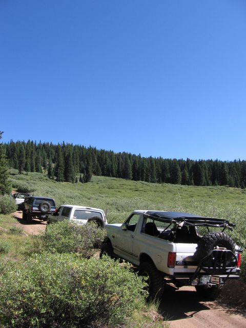 1995 Ford Bronco - Several off-road vehicles, including a white Ford Bronco, navigate a dirt path through a grassy, wooded area.