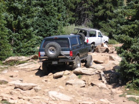 1995 Ford Bronco - Two Ford Broncos navigate a challenging rocky trail in a wooded mountain area.