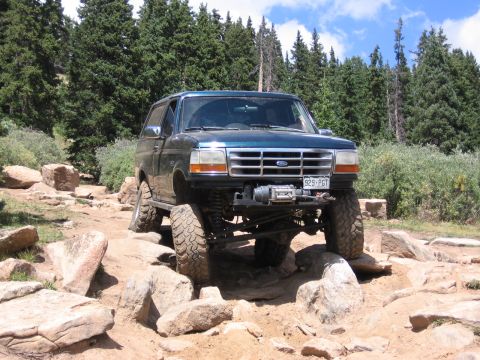 1995 Ford Bronco - A teal 1995 Ford Bronco tackles a challenging rocky path in a wooded mountain area, its off-road capabilities.