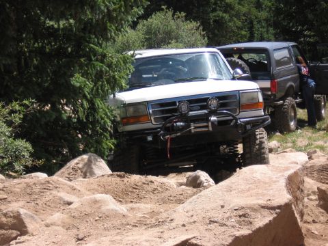 1995 Ford Bronco - Two Ford Broncos navigate a challenging off-road course with large rocks and dirt mounds.