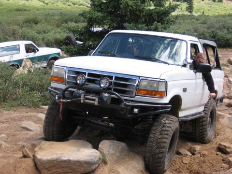 1995 Ford Bronco - Two Ford Broncos tackle a challenging off-road trail with large rocks and dense vegetation.