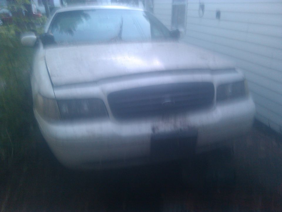 Front view of a white Ford Crown Victoria sedan parked next to a white-sided house