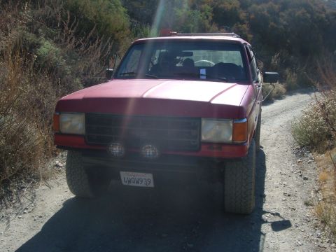 1988 Ford Bronco - Faded red full-size Ford Bronco photographed head-on while stopped on a narrow dirt fire road cut into brushy hills. It has a pair of round…