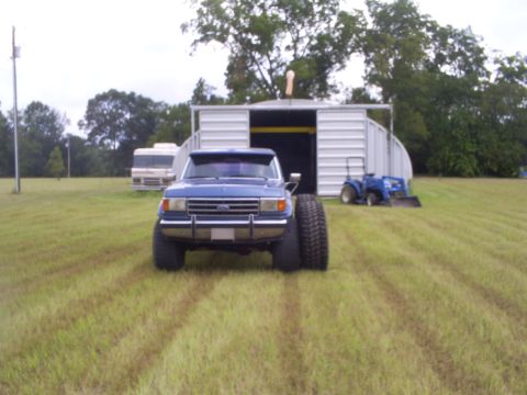 1992 Ford Bronco - A blue pickup truck with massive tires is parked in a grassy field in front of a metal building.